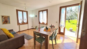 Una sala de estar con un sofá, una mesa de comedor con sillas, una lámpara de pie y vistas a un jardín a través de una puerta.