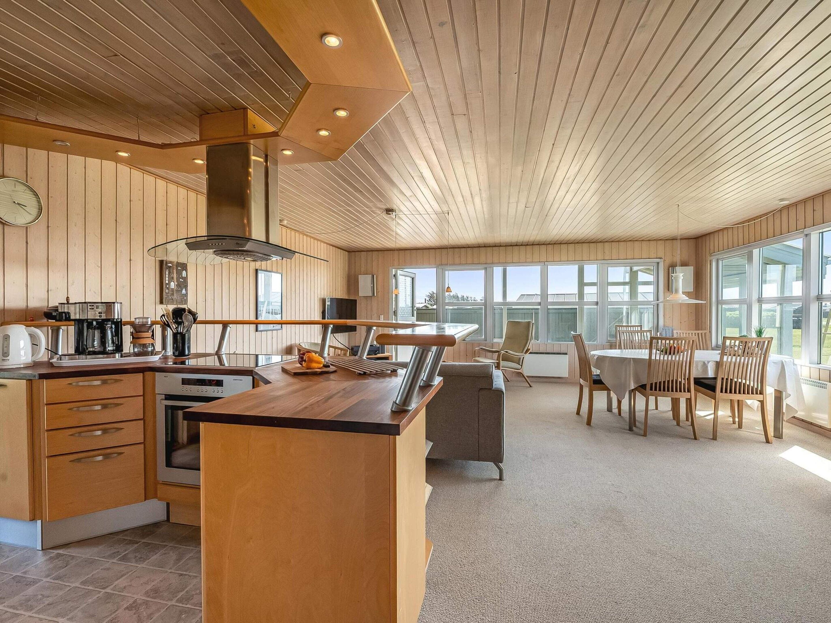 A modern kitchen with wooden paneling, a dining area with a table and chairs, and large windows providing natural light.