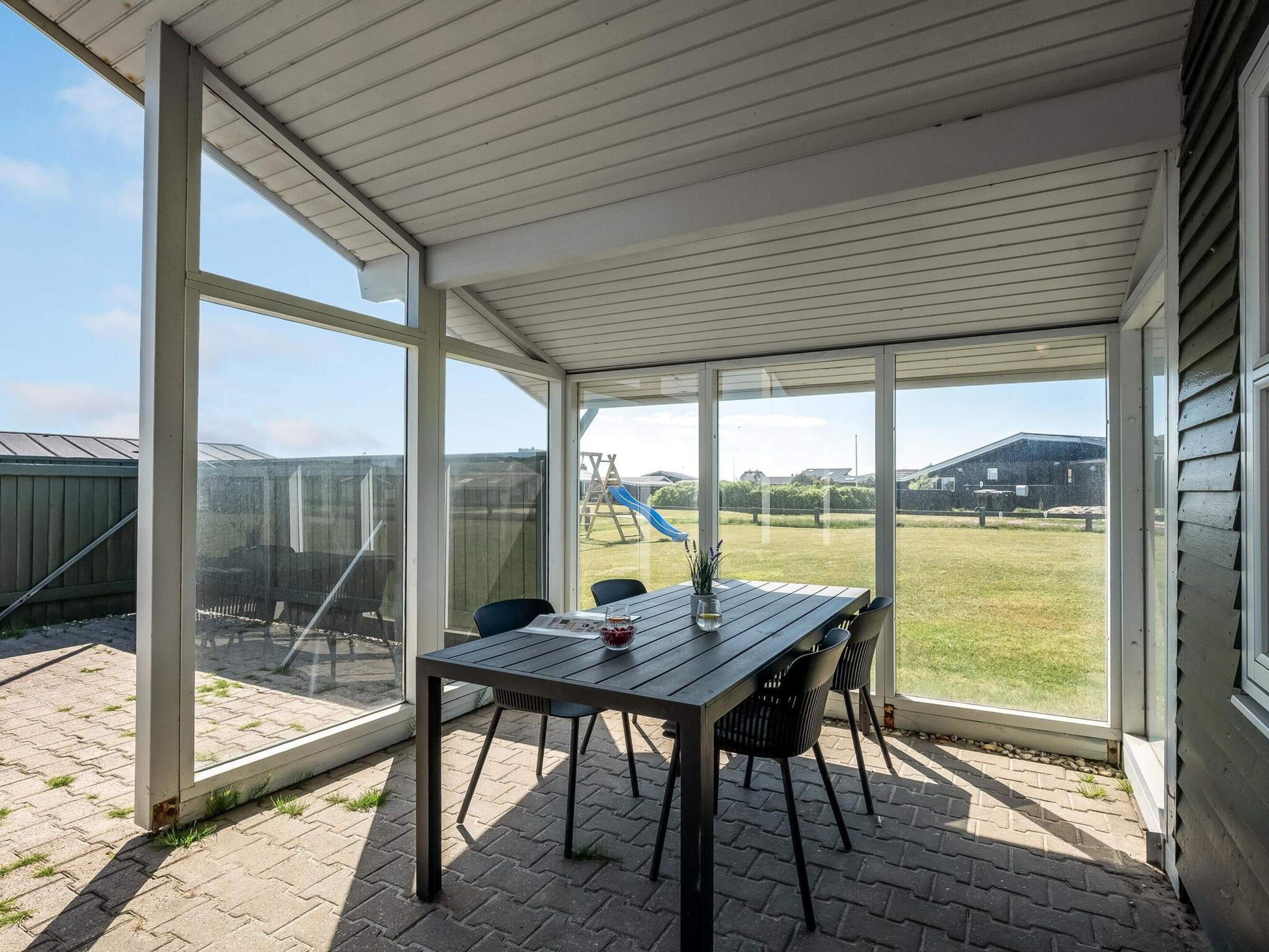 Une terrasse couverte avec une table et des chaises, de grandes fenêtres et une vue sur une aire de jeux.