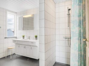 Ein Badezimmer mit weißen Fliesen, einer Dusche mit Glastür, einem Waschbecken mit zwei Pflanzen und einem Fenster mit Milchglas.