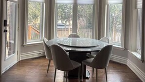 Un coin repas comprenant une table ronde, quatre chaises, un lustre et de grandes fenêtres équipées de stores.