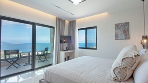 Ein Schlafzimmer mit einem großen Bett, einem Schreibtisch und Meerblick durch raumhohe Fenster.