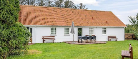 A white building with a red-tiled roof, a patio with a table and chairs, and a grassy yard.
