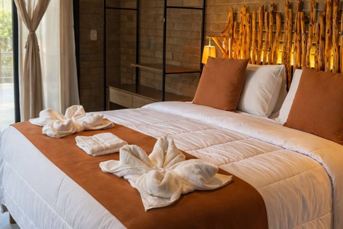 Ein Hotelzimmer mit Backsteinwand, einem Bett mit weißer Bettwäsche und braunen Akzenten, einem Holz-Kopfteil und einem großen Fenster mit Vorhängen.