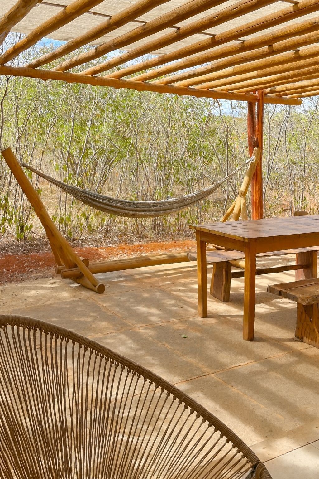 Una hamaca bajo una pérgola de madera, una mesa de madera y una silla de mimbre en un entorno al aire libre con árboles al fondo.