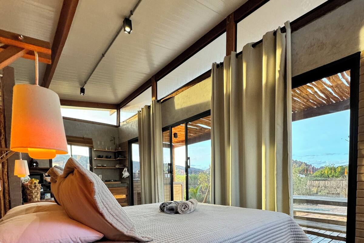 Ein Schlafzimmer mit einem großen Bett, einem hölzernen Kopfteil, einer Nachttischlampe und Blick ins Freie durch große Fenster.