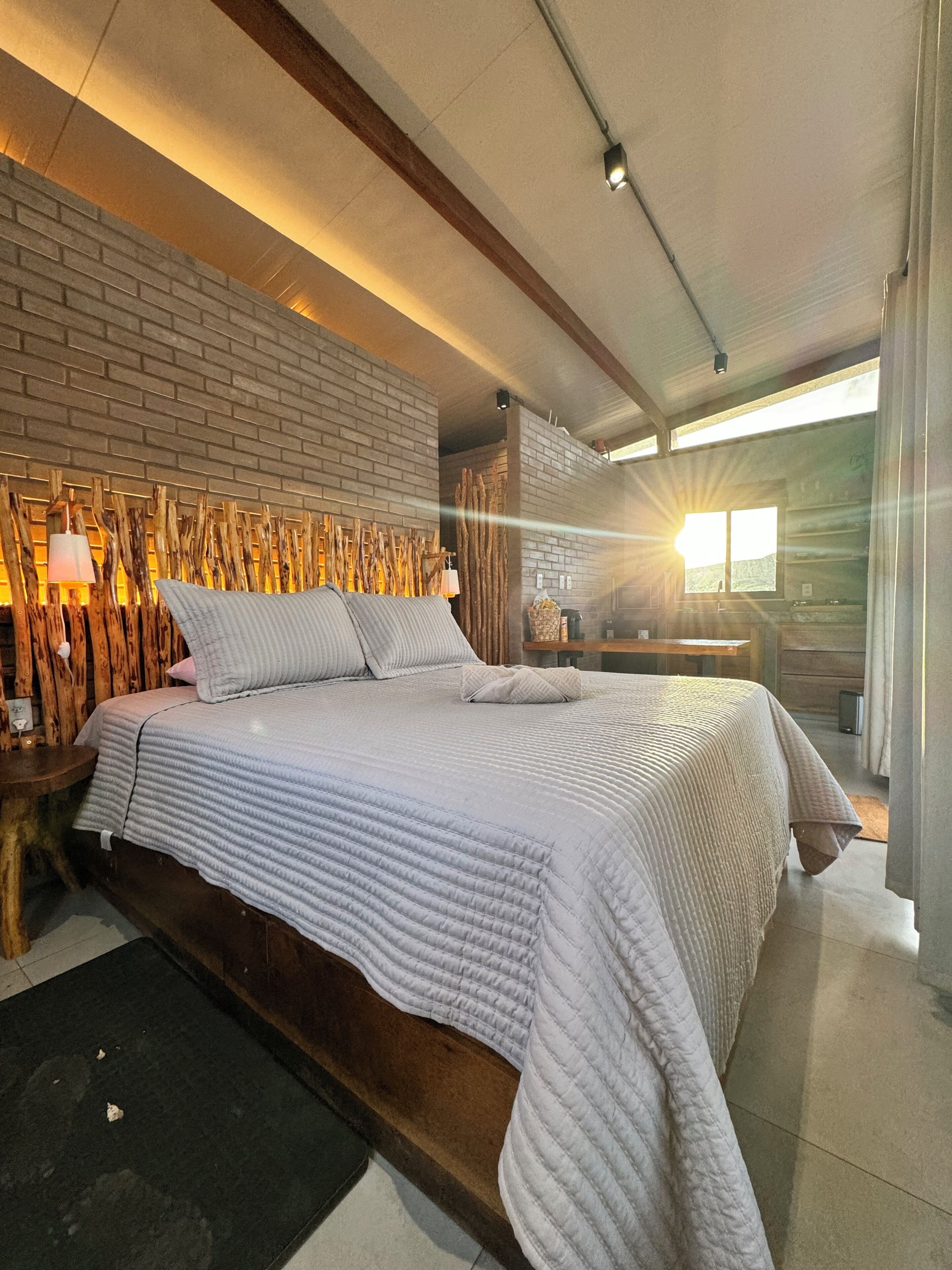 A modern bedroom with a brick wall, a large bed with a quilted cover, wooden headboard, and a nightstand with a lamp.