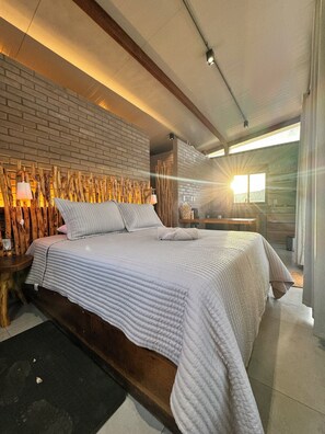 A modern bedroom with a brick wall, a large bed with a quilted cover, wooden headboard, and a nightstand with a lamp. - Sertão do luar (Coronel José Dias)