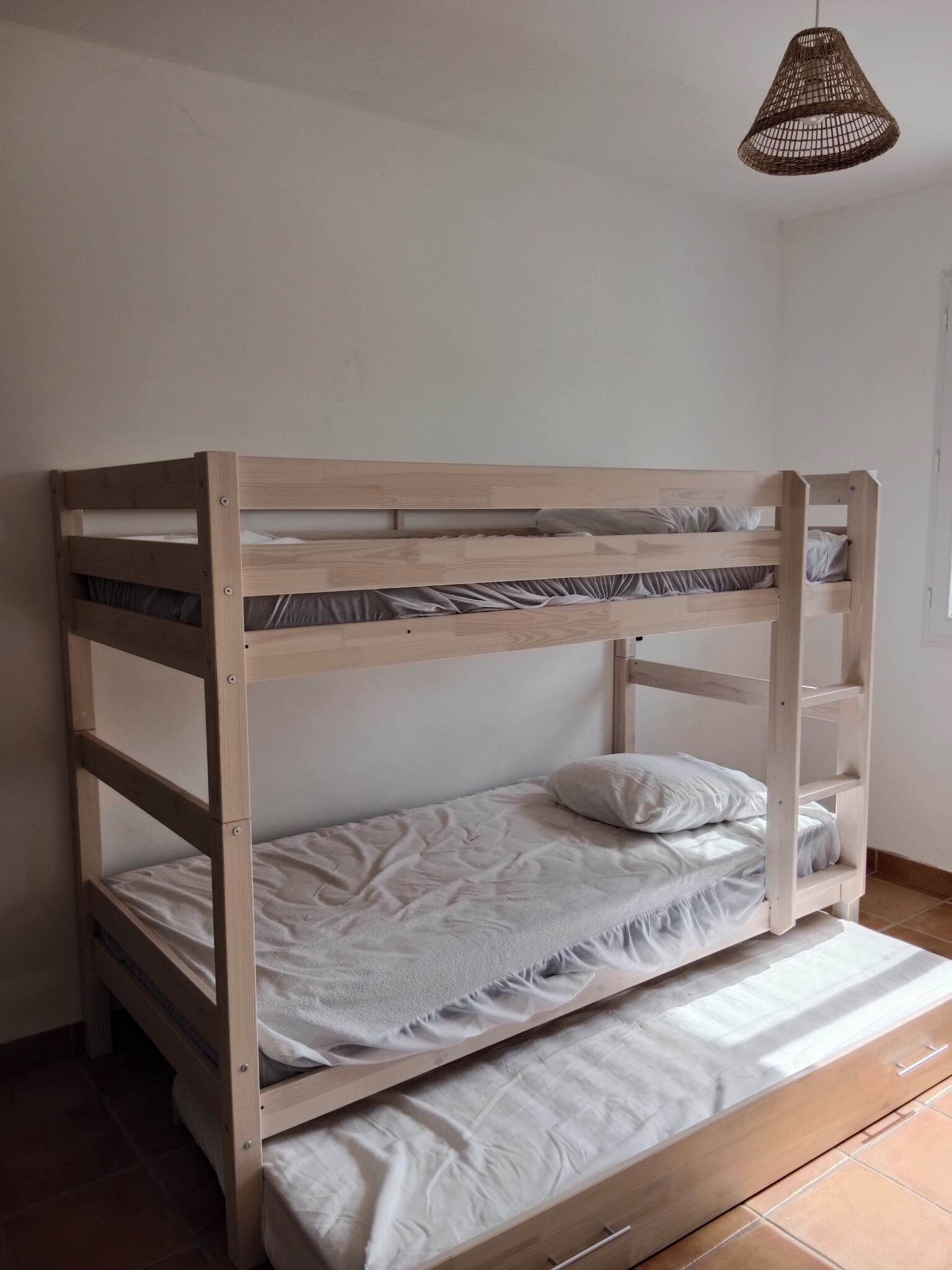 Een stapelbed met houten frame, een enkel matras in het onderste bed, en een hanglamp erboven.