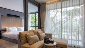 Ein modernes Hotelzimmer mit Bett, Sofa, Hocker, einem runden Beistelltisch und großen Fenstern mit Vorhängen.