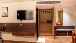 A hotel room with a wooden bench, a television, a coat rack, and a bathroom with a shower curtain. - Hotel Golden Amulya Rest Wings (Bengaluru)