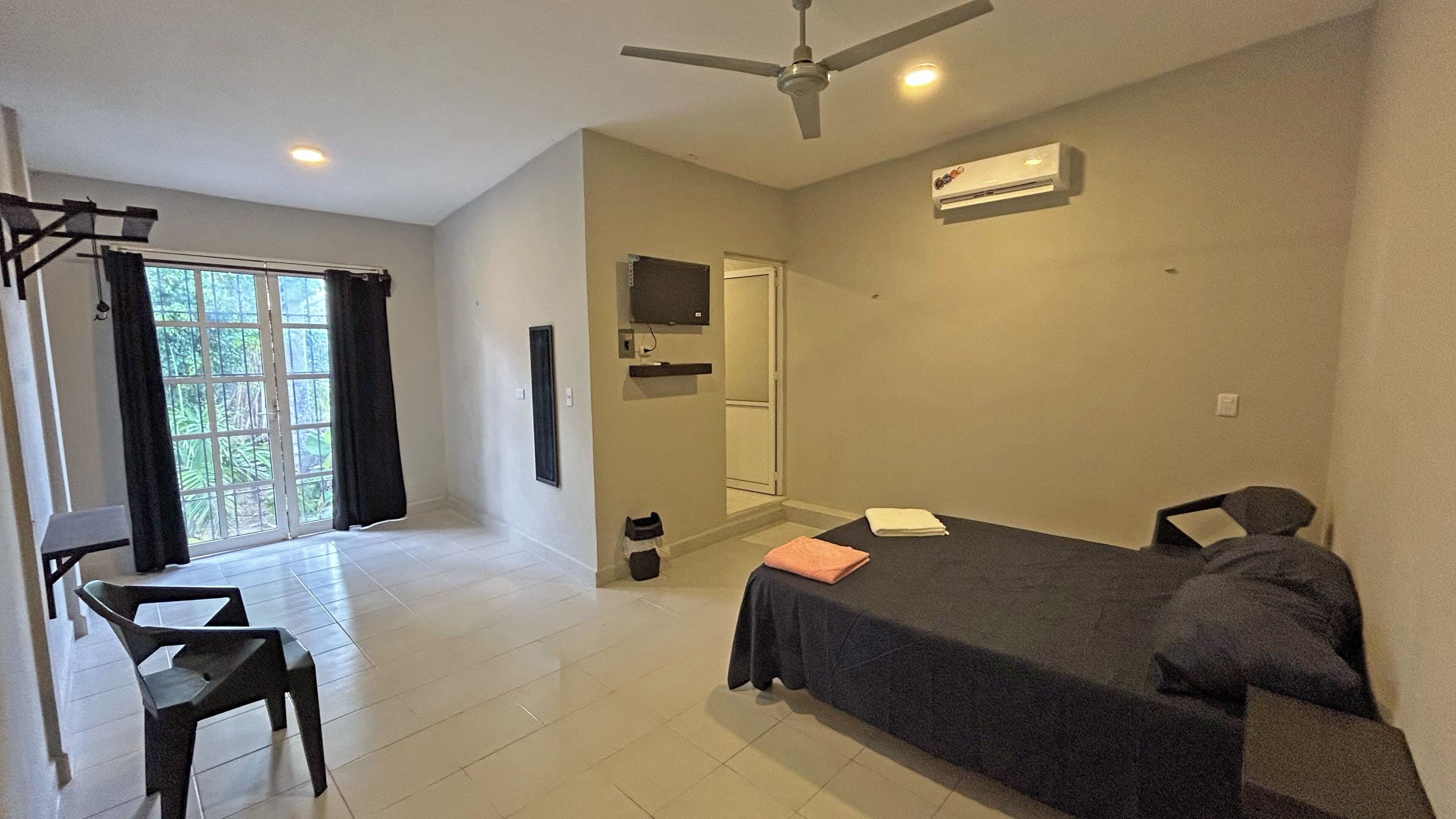A bedroom with a bed, a desk, a chair, a ceiling fan, a TV, and a window with curtains.