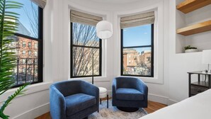 A corner room with two blue armchairs, a white sofa, a round rug, and a view of buildings through large windows. - Cityscape 1BR Plus: King + Queen Pull-out | Suite 7 (Boston)