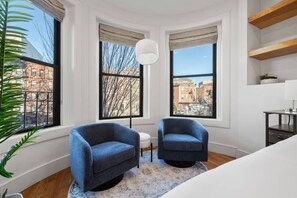 Ein Eckzimmer mit zwei blauen Sesseln, einem weißen Sofa, einem runden Teppich und Blick auf Gebäude durch große Fenster.