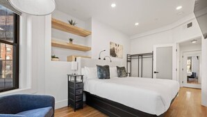 A modern bedroom with a large bed, built-in shelves, a bedside table, and a chair. - Cityscape 1BR Plus: King + Queen Pull-out | Suite 7 (Boston)