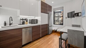 A modern kitchen with white cabinets, a dishwasher, a refrigerator, and a dining area with a table and chairs. - Cityscape 1BR Plus: King + Queen Pull-out | Suite 7 (Boston)