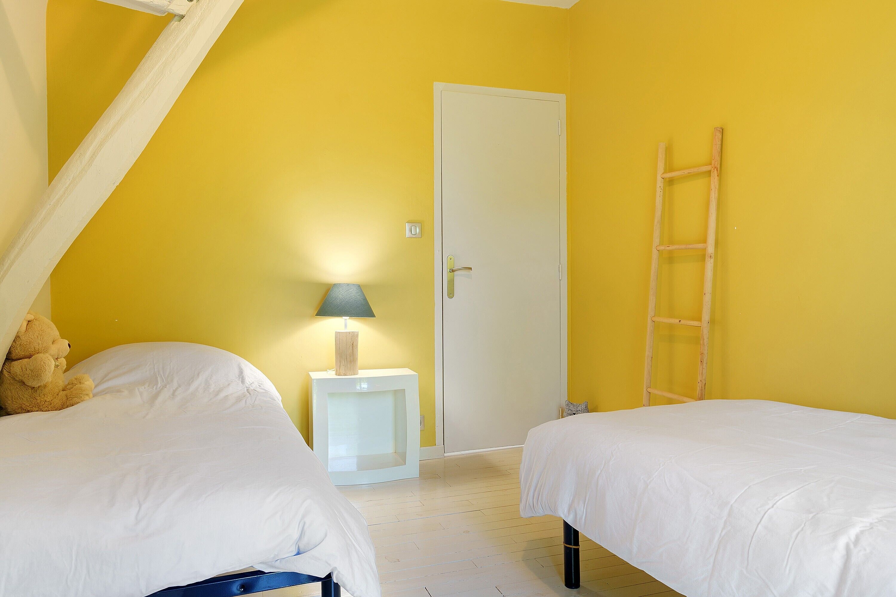 Ein Zimmer mit zwei Betten, einer weißen Tür, einem kleinen Nachttisch mit Lampe und einer Holzleiter an der Wand.