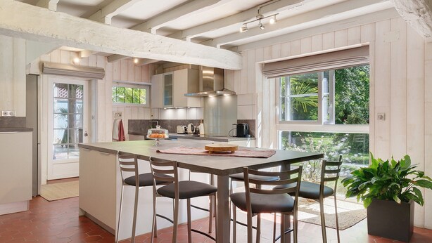 A kitchen with a central island, bar stools, a potted plant, and a view through the window.