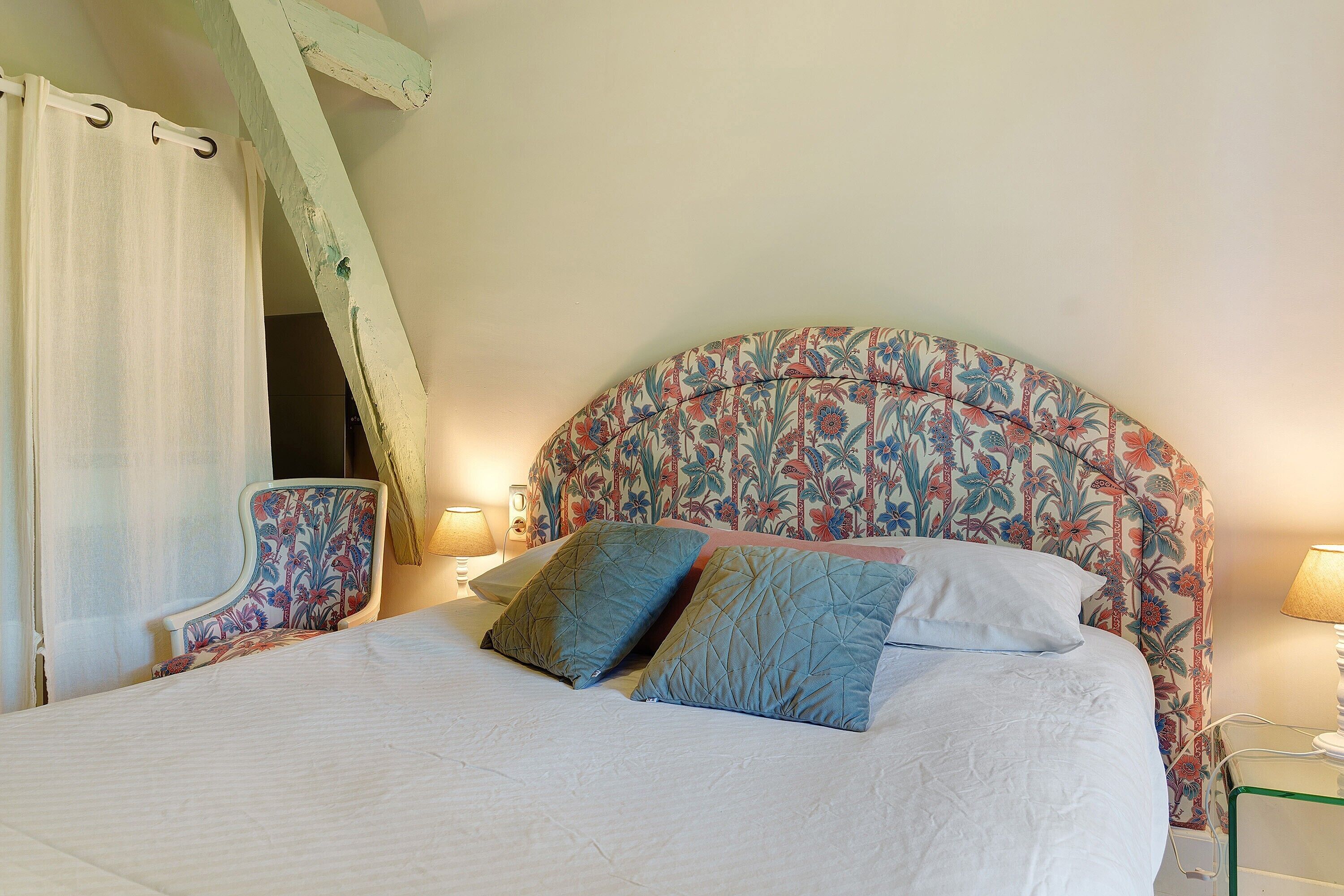 A bed with a floral headboard, a chair with a floral pattern, and a bedside table with a lamp.