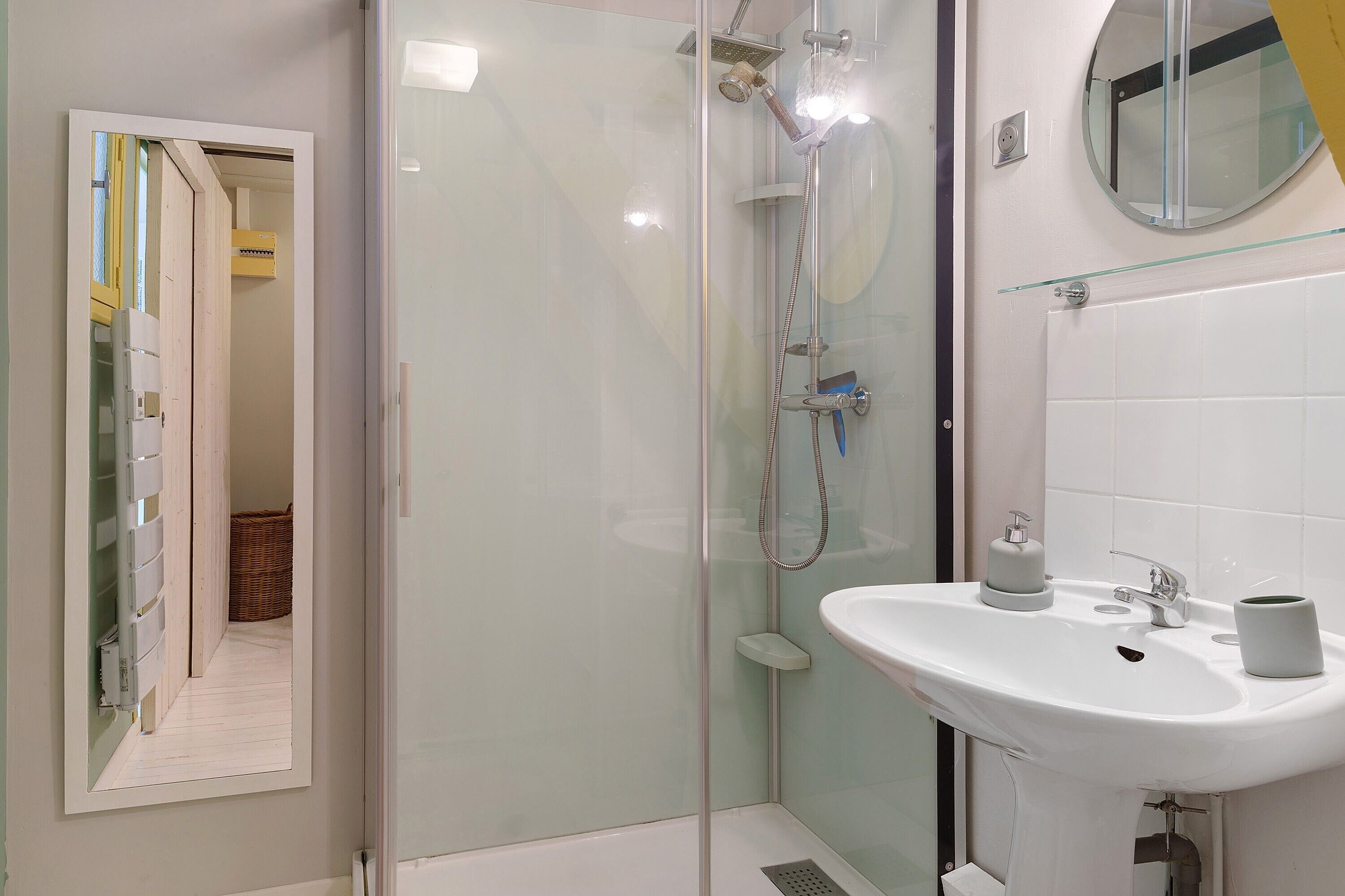 Een badkamer met een douchecabine van glas, een witte wastafel met spiegel erboven en een deur die naar een andere kamer leidt.