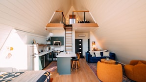 A compact, modern interior with a loft bedroom, kitchenette, and living area, featuring wooden floors and a central island. - Oak Hollow | Hocking Hills, Hot Tub, Romantic (McArthur)