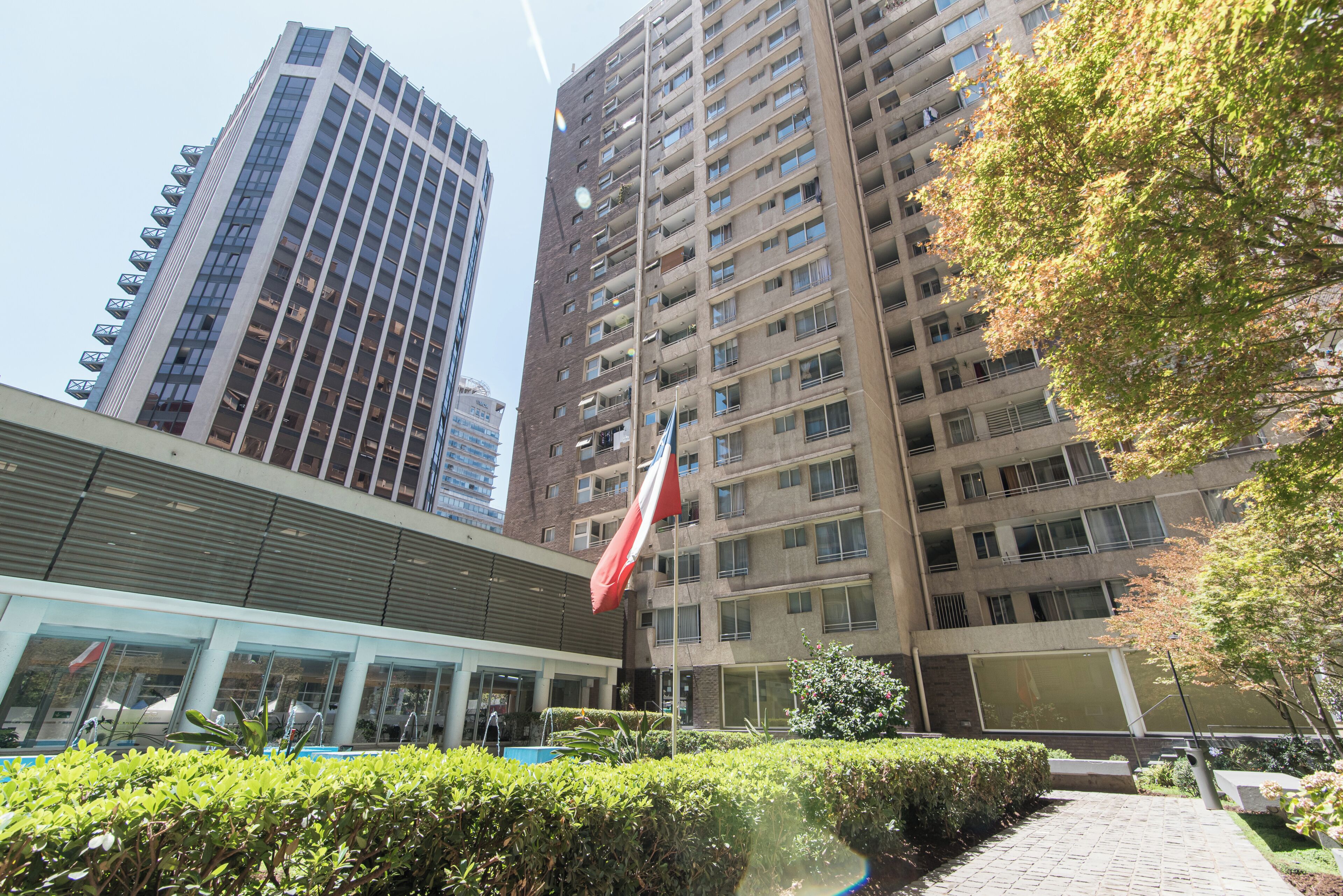 Un edificio de varios pisos con una bandera, fachada de vidrio y zona ajardinada con arbustos.