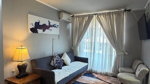 Een moderne woonkamer met een bank, een bijzettafeltje met een lamp, een kamerkleed met patroon, en een groot raam met gordijnen.
