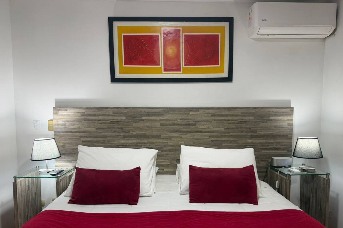 A hotel room with a wooden headboard, two bedside tables with lamps, a bed with white and red bedding, and a framed artwork above the bed.