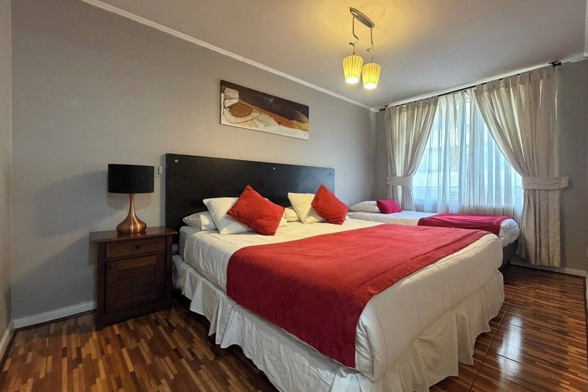 A hotel room with two beds, a nightstand, a lamp, a painting on the wall, and a window with curtains.