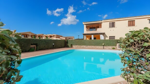 Una piscina con cielo azul despejado, rodeada de edificios con balcones y un seto.