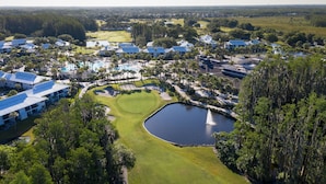 An aerial view of a golf course with a pond, surrounded by buildings with blue roofs, and dense greenery. - Saddlebrook Resort | 1BR 2 Queen | Tennis Programs, Golf Courses, Nature Trails (Wesley Chapel)