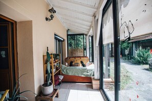 Ein Zimmer mit einem Bett, einem hölzernen Regal mit Pflanzen, einem Kakteen im Topf und einem großen Fenster.
