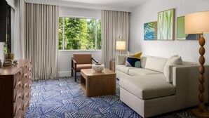 Ein Hotelzimmer mit einer Couch, einem Sessel, einem Holztischchen, einer Lampe und einem Fenster mit Vorhängen.