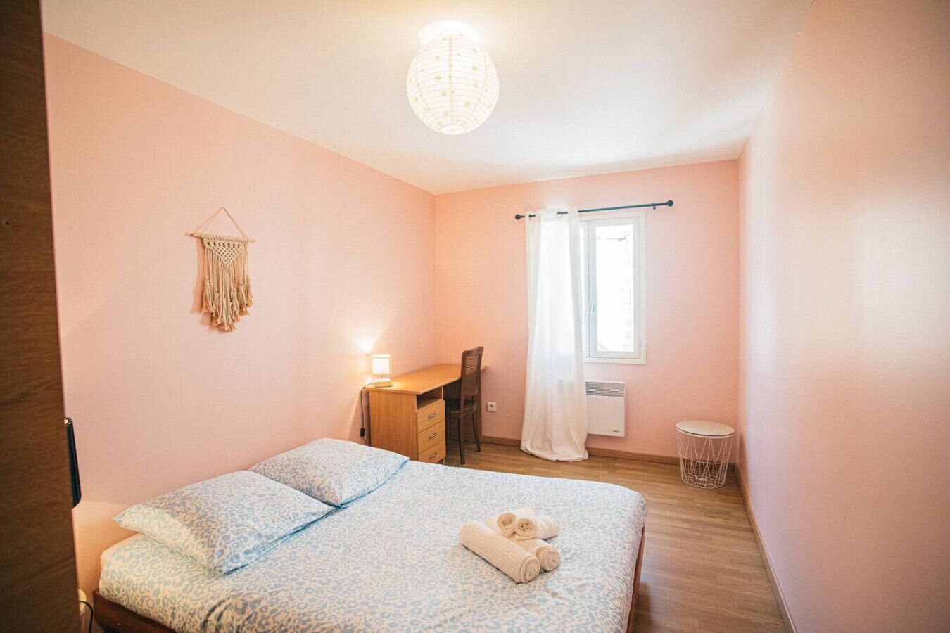 Un dormitorio con cama, escritorio, silla, una mesita redonda, un tapiz de macramé y una ventana con cortinas.