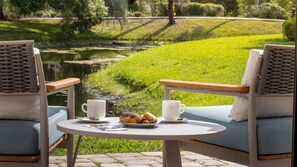 Zwei Stühle mit gewobenen Rückenlehnen und hellblauen Kissen, ein kleiner runder Tisch mit einer weißen Tasse und einem Teller mit Essen, und Blick auf einen Teich und eine Rasenfläche.
