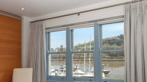 Quarto com uma grande janela, uma cama, uma cadeira e vista para barcos no rio.