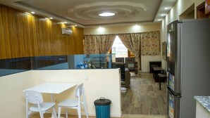 A compact kitchen with a refrigerator, a dining table with chairs, and a trash bin. - RESIDENCE MBESSA (Abidjan)