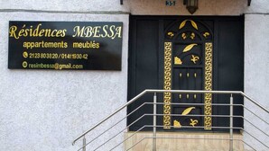 A building facade with a black and gold door, a signboard with contact information, and a metal staircase. - RESIDENCE MBESSA (Abidjan)
