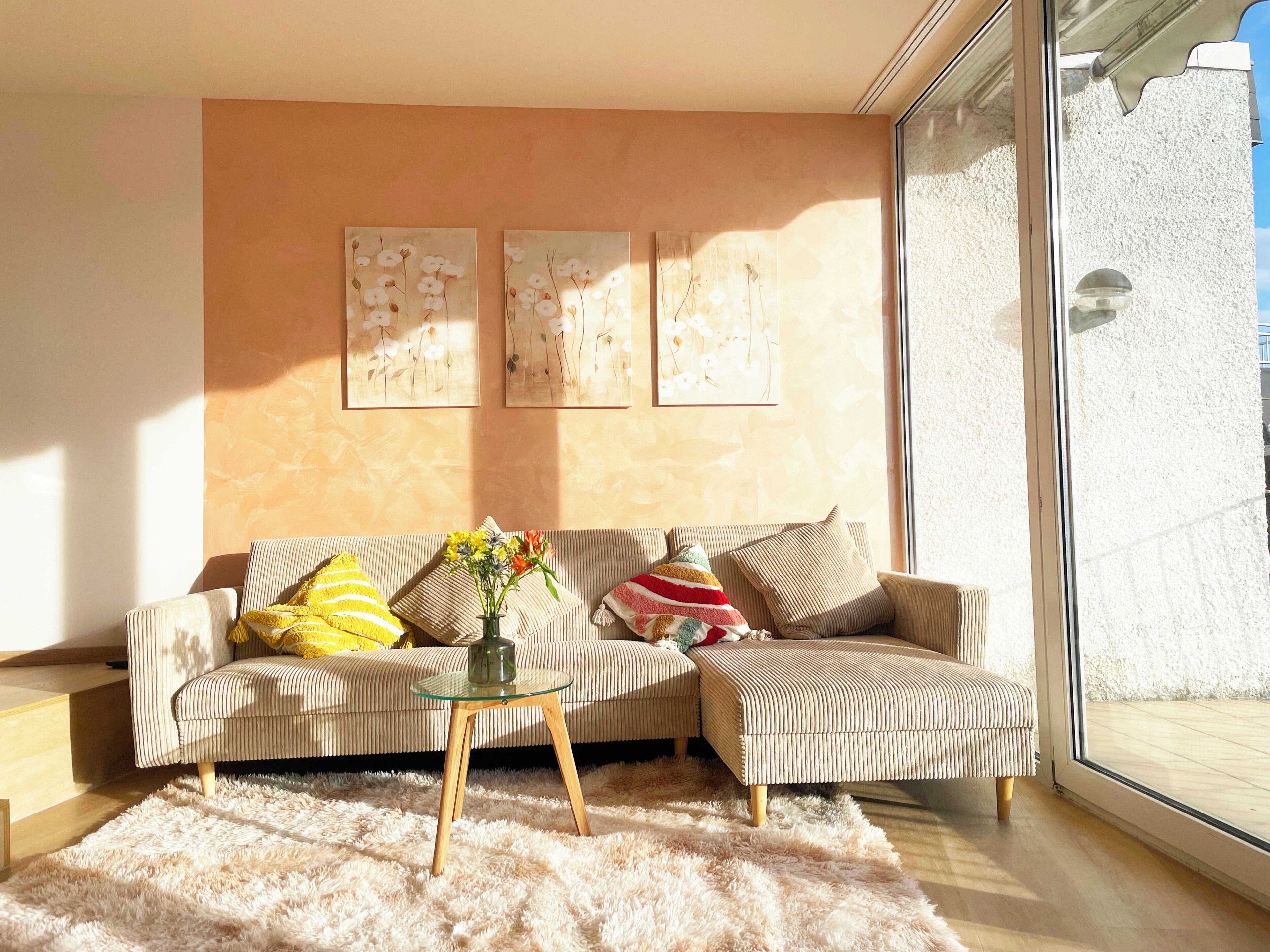 A living room with a sofa, ottoman, glass coffee table, and a rug. There are three wall-mounted paintings and a large window.