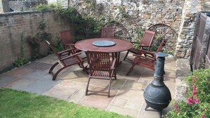 A patio with a round table and chairs, a black outdoor heater, and a stone wall with vegetation. - Near Beach - Pets - Bbq - Fireplace - Sleeps 10 (Leven)