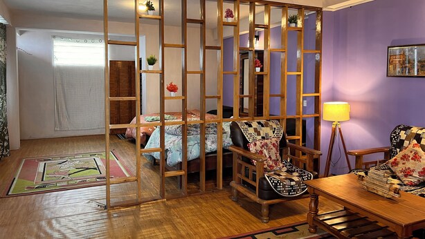 A room with a wooden partition, a bed with floral bedding, a small table with a lamp, and a decorative pillow.