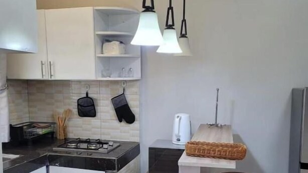 A kitchen with white cabinets, a black countertop, a gas stove, hanging kitchen utensils, a coffee maker, and a bar stool.