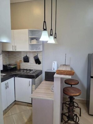 A kitchen with white cabinets, a black countertop, a gas stove, hanging kitchen utensils, a coffee maker, and a bar stool.