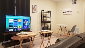 Ein Wohnzimmer mit Fernseher, der eine Streaming-Dienstanwendung anzeigt, einem Holztisch mit einem Weinglas, einem Sofa und einem kleinen runden Tisch mit einem Stuhl.