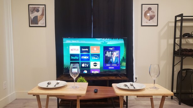 Ein Wohnzimmer mit Fernseher, der Streaming-Dienste anzeigt, zwei Esstischen mit Tellern und Besteck sowie einem dunklen Vorhang.