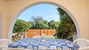 Eine Terrasss mit einem Tisch, der mit einer blau-blumigen Tischdecke bedeckt ist, umgeben von weißen Plastikstühlen.