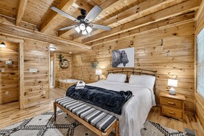 Un dormitori de cabana de fusta amb un llit, taules de nit, un ventilador de sostre i un quadre penjat a la paret.