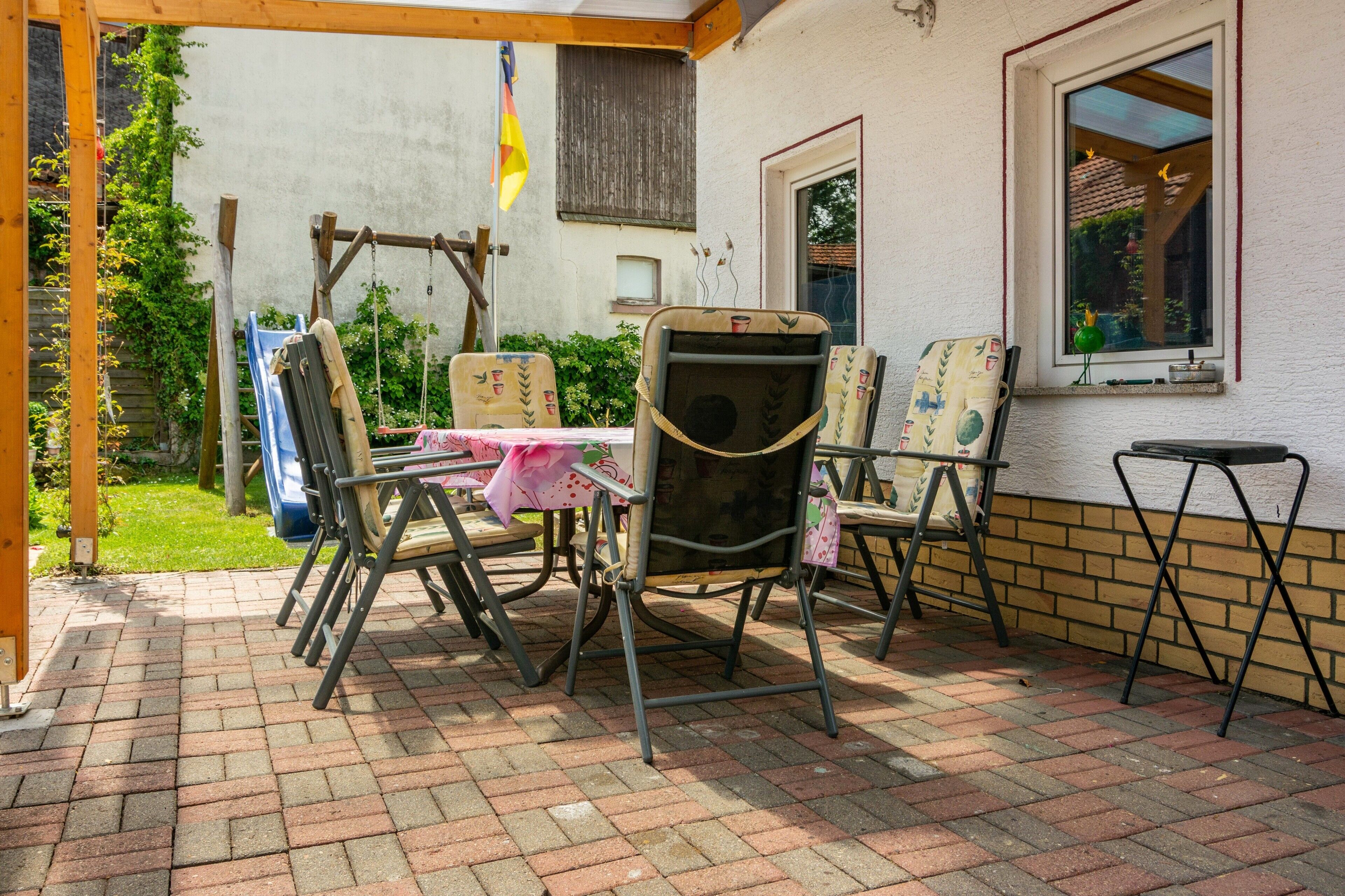 En patio med bord og stole, et gyngestativ og et vindue med udsigt til grønne omgivelser.