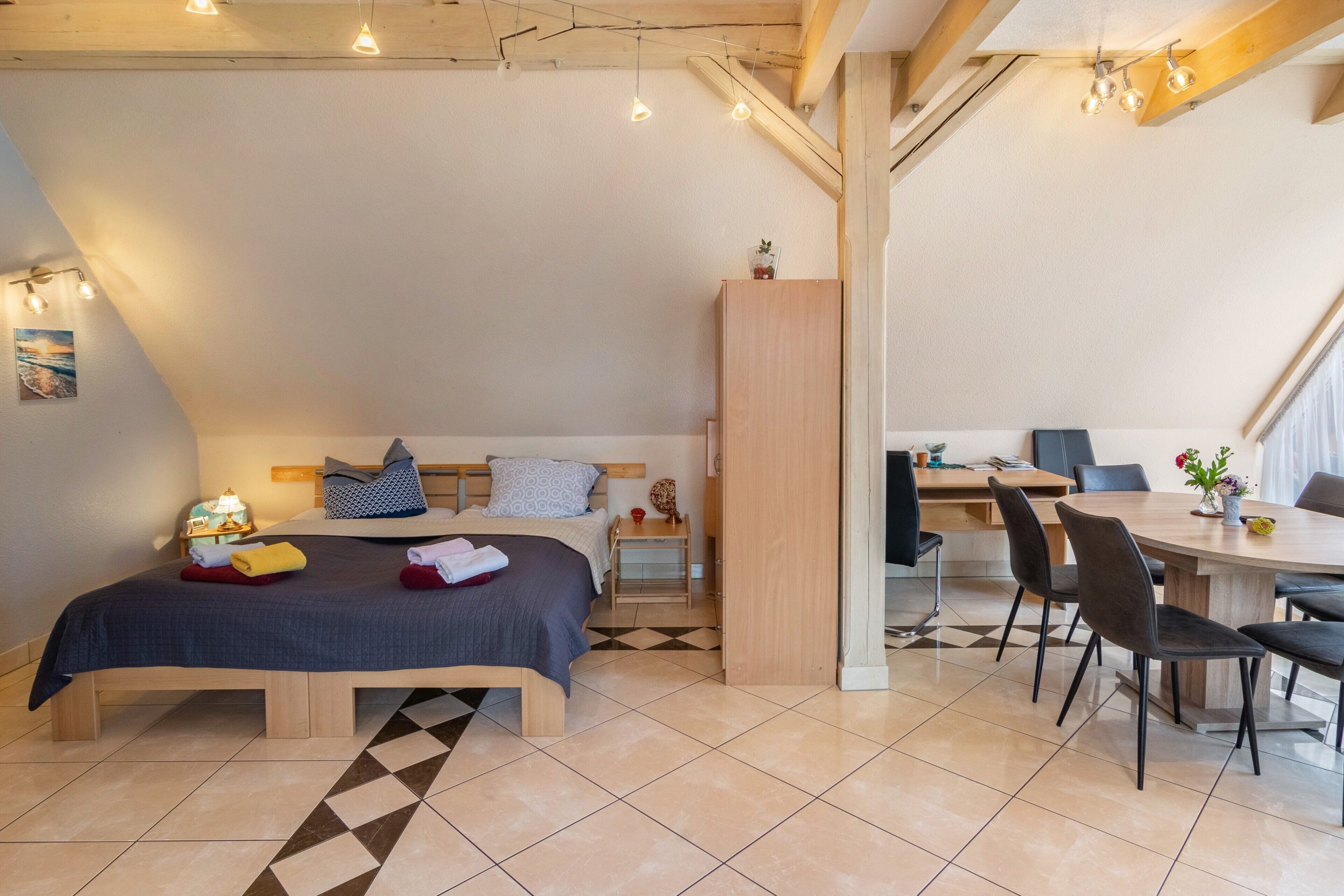 A bedroom with a bed, bedside table, and a small cabinet. There is a dining area with a table and chairs.