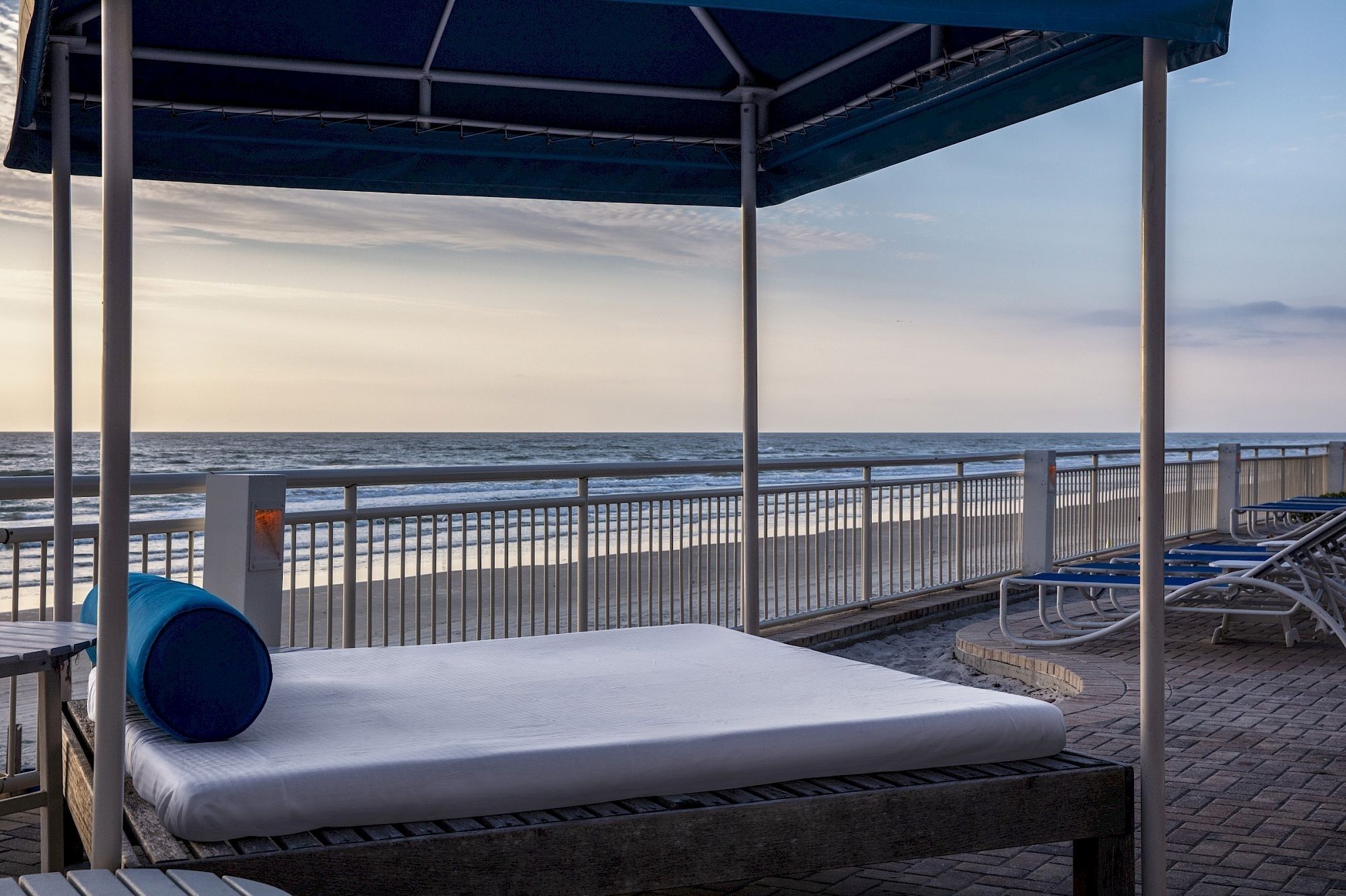 A beachside lounge with a covered seating area, a view of the ocean, and a metal railing.